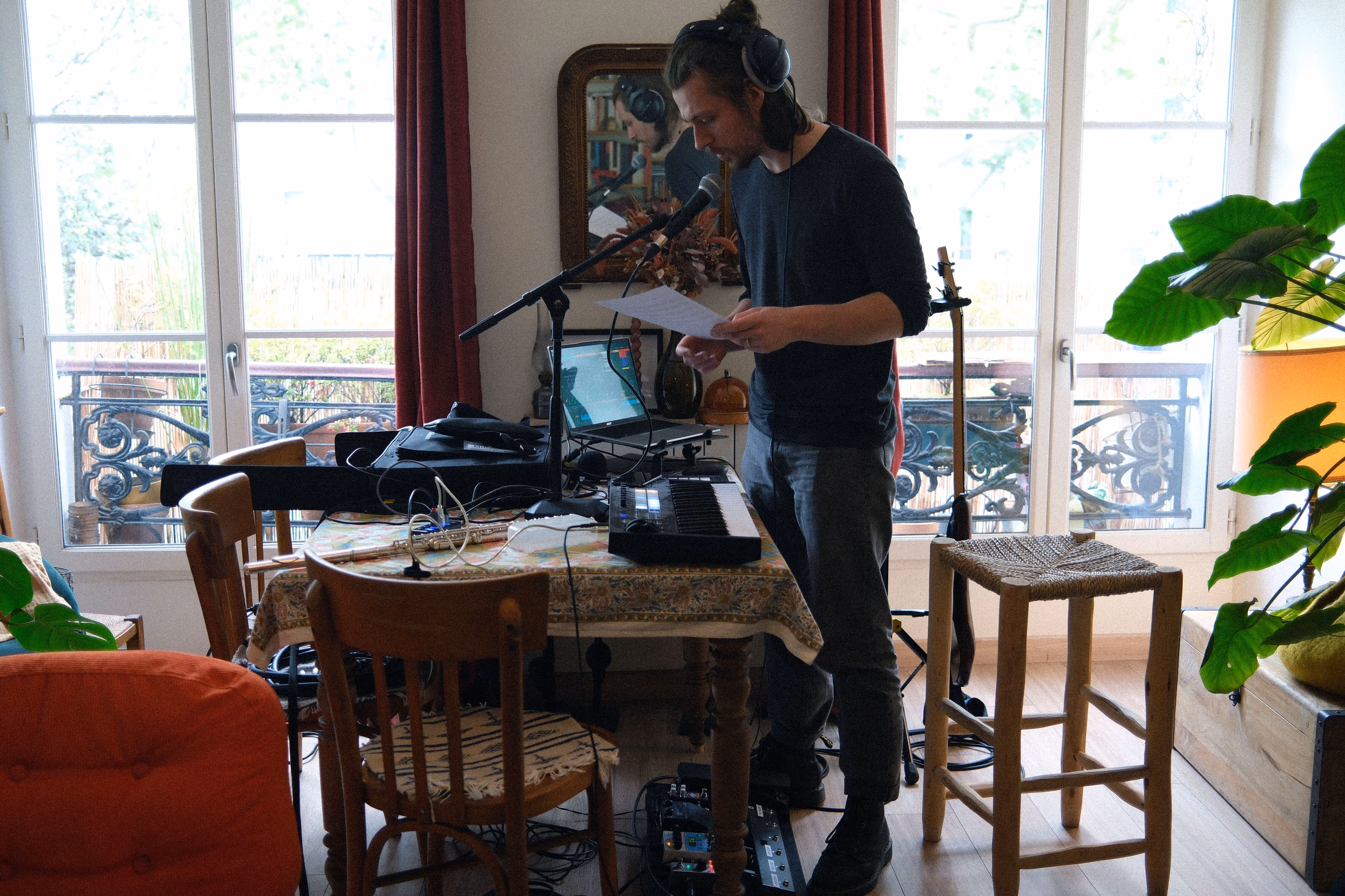 Un homme avec un casque audio lit une feuille devant un micro, entouré d'un clavier MIDI et d'un ordinateur portable dans un salon aménagé en studio