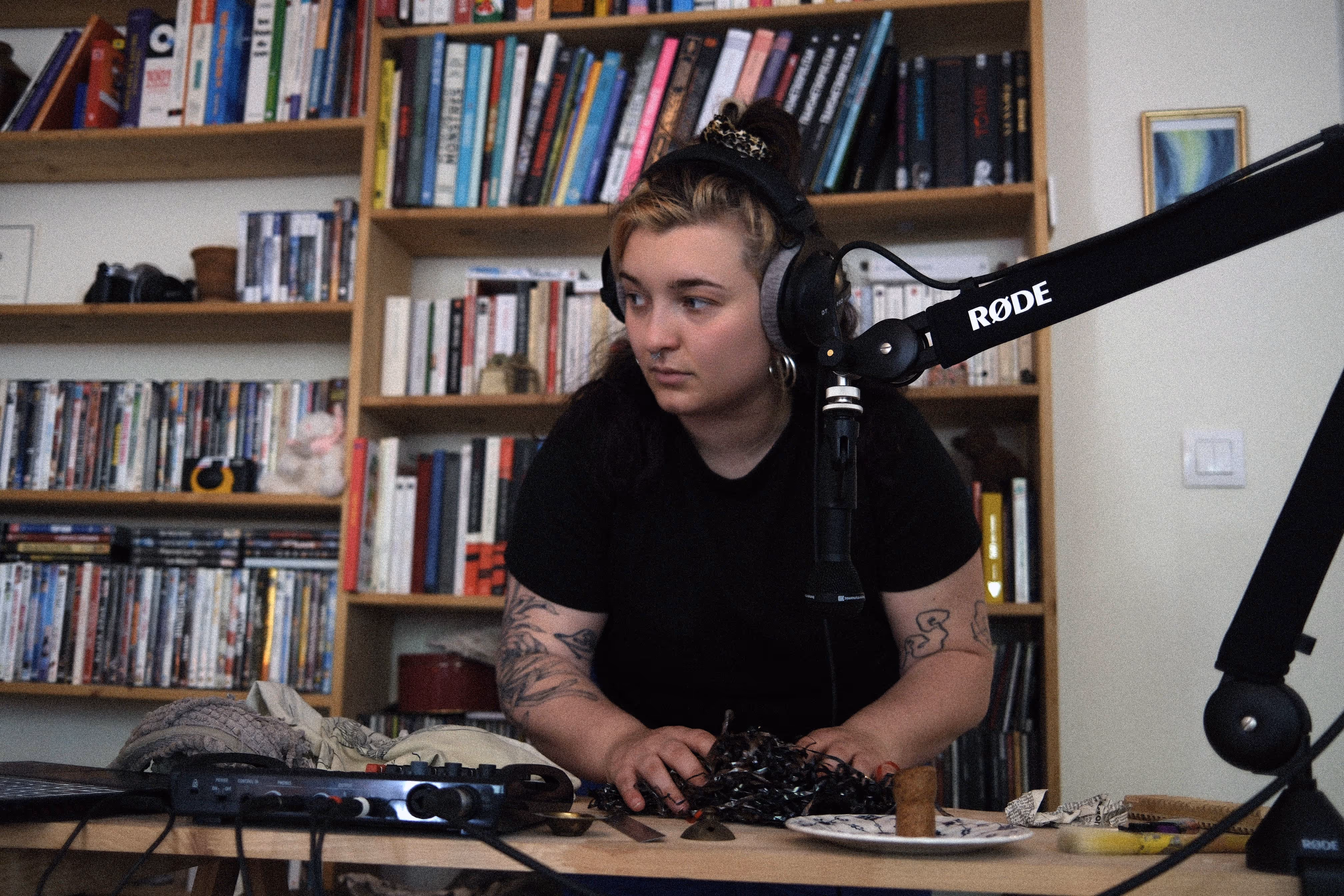 Une femme avec un casque audio manipule des objets devant un micro Røde, dans une pièce avec une bibliothèque remplie de livres et DVD en arrière-plan